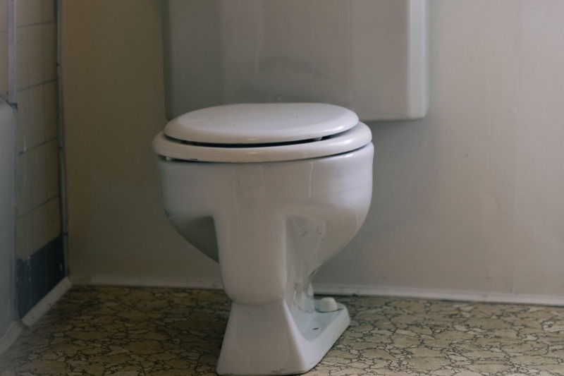 white toilet with a closed seat against a wall in an older bathroom with a patterned floor